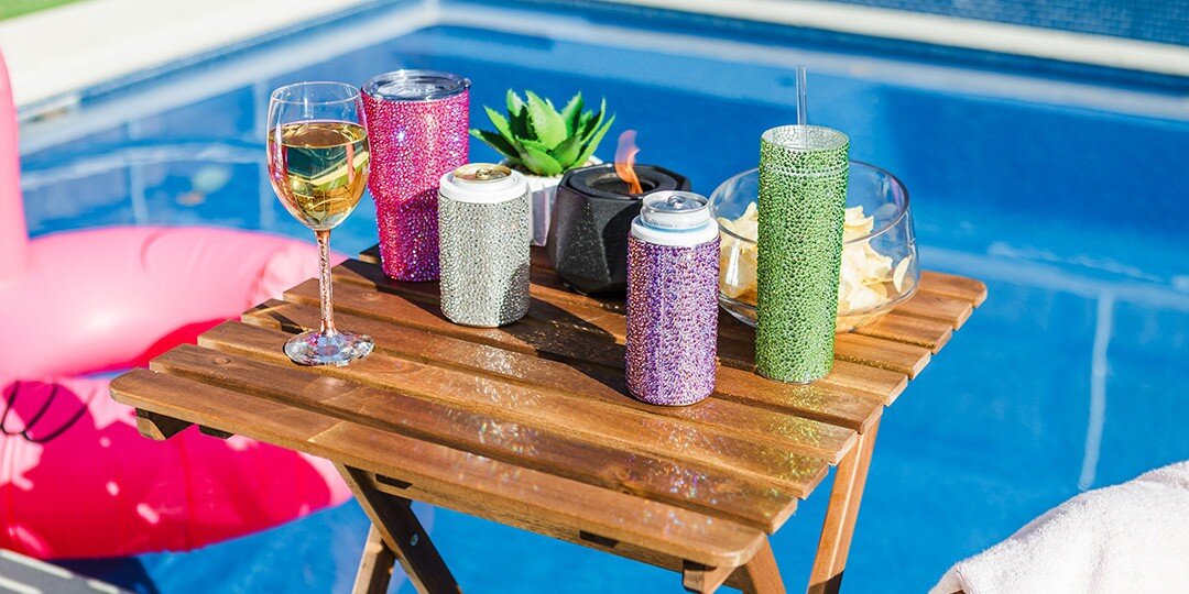 Poolside&amp;&#x23;x20&#x3B;wooden&amp;&#x23;x20&#x3B;table&amp;&#x23;x20&#x3B;displaying&amp;&#x23;x20&#x3B;collection&amp;&#x23;x20&#x3B;of&amp;&#x23;x20&#x3B;rhinestone&amp;&#x23;x20&#x3B;drink&amp;&#x23;x20&#x3B;accessories&amp;&#x23;x20&#x3B;including&amp;&#x23;x20&#x3B;pink,&amp;&#x23;x20&#x3B;silver,&amp;&#x23;x20&#x3B;green,&amp;&#x23;x20&#x3B;and&amp;&#x23;x20&#x3B;purple&amp;&#x23;x20&#x3B;sparkly&amp;&#x23;x20&#x3B;can&amp;&#x23;x20&#x3B;coolers&amp;&#x23;x20&#x3B;and&amp;&#x23;x20&#x3B;tumblers,&amp;&#x23;x20&#x3B;wine&amp;&#x23;x20&#x3B;glass,&amp;&#x23;x20&#x3B;and&amp;&#x23;x20&#x3B;small&amp;&#x23;x20&#x3B;succulent&amp;&#x23;x20&#x3B;plant,&amp;&#x23;x20&#x3B;with&amp;&#x23;x20&#x3B;blue&amp;&#x23;x20&#x3B;swimming&amp;&#x23;x20&#x3B;pool&amp;&#x23;x20&#x3B;and&amp;&#x23;x20&#x3B;pink&amp;&#x23;x20&#x3B;float&amp;&#x23;x20&#x3B;visible&amp;&#x23;x20&#x3B;in&amp;&#x23;x20&#x3B;background.