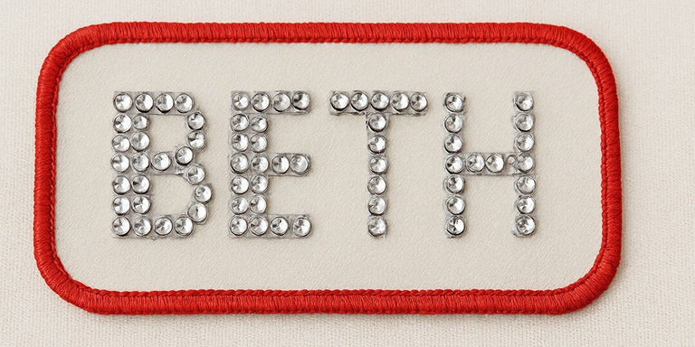 Un parche de nombre decorativo con el nombre BETH escrito en estrás plateados en un fondo de tela beige con un borde rojo brillante bordado