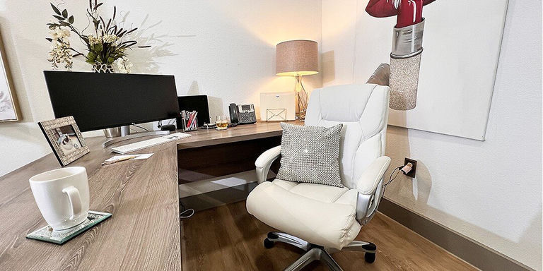 A modern home office featuring a light wood desk with dual black monitors, a white ergonomic office chair with a silver mirrored rhinestone throw pillow, and a warm table lamp with a beige shade. The workspace includes office supplies, a decorative plant arrangement, and contemporary artwork on white walls. Dark hardwood floors complete the clean, professional aesthetic.