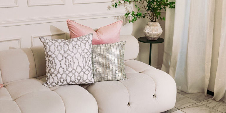 White sectional sofa decorated with blush pink velvet pillows, silver mirrored rhinestone throw pillow, and white pillow with a silver lattice pattern. Black side table holds textured vase with greenery, set against white paneled wall with flowing curtains.