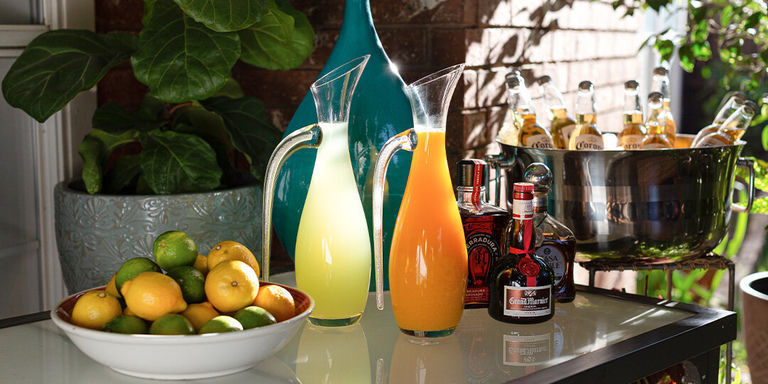Un chariot de bar sophistiqué présentant un assortiment de cocktails et de spiritueux. Divers verres contiennent des boissons de couleurs différentes, dont du vin rouge et blanc, un cocktail ambré dans une coupe et une boisson foncée. Des bouteilles vertes de vin ou de champagne, un shaker à cocktail argenté et une flasque étincelante incrustée de strass sont disposés sur le chariot. Un vase blanc côtelé contient une plante décorative avec de petites oranges, et un fond géométrique blanc à motifs complète la mise en scène.