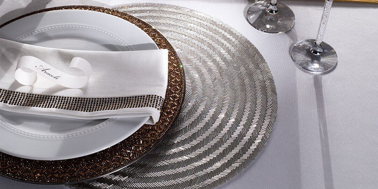 Mesa con plato blanco sobre cargador de bronce con pedrería, colocado sobre mantel individual plateado con patrón de chevron. Servilleta blanca con anillo de pedrería y tarjeta de lugar pequeña dispuestos junto a cristalería en mantel blanco.