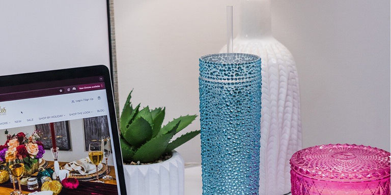 Espacio de trabajo en escritorio con laptop mostrando sitio web, vaso azul con diamantes de imitación y pajita, pequeña planta suculenta en maceta blanca y contenedor rosa con diamantes de imitación, con jarrón blanco texturizado en el fondo.