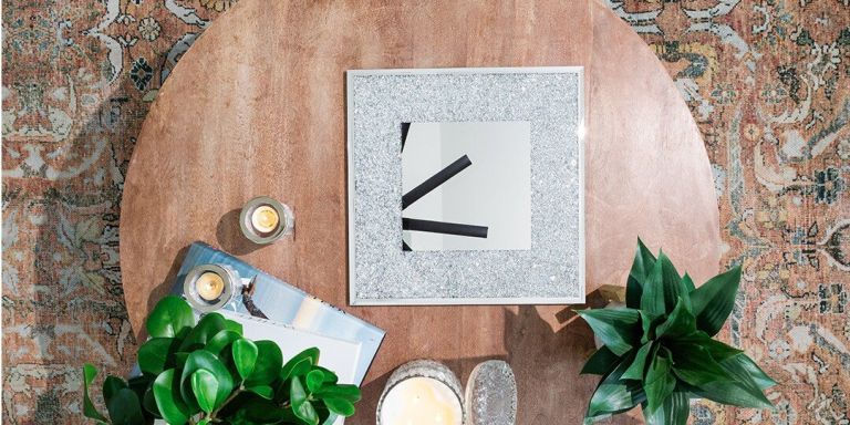 Una vista aérea de una mesa de café redonda de madera decorada con accesorios decorativos, colocada sobre una alfombra vintage con patrón en tonos tierra. La mesa cuenta con una bandeja cuadrada con espejo bordeada de pequeños diamantes de plata bajo el cristal, flanqueada por plantas en macetas verdes, pequeñas velas encendidas en portavelas de vidrio y un libro. La composición crea una viñeta de espacio de vida moderno y curado.