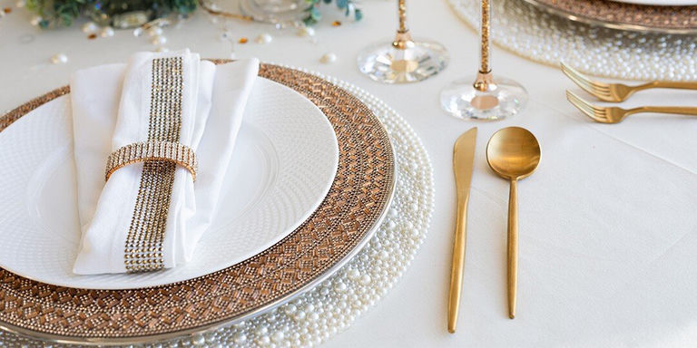Mise de table luxueuse avec assiette plate blanche sur assiette de présentation en strass bronze, superposée à un set de table blanc à perles. Serviette blanche maintenue par un lien doré en strass, accompagnée de couverts dorés et de verres en cristal sur nappe blanche, avec des touches de verdure en arrière-plan.