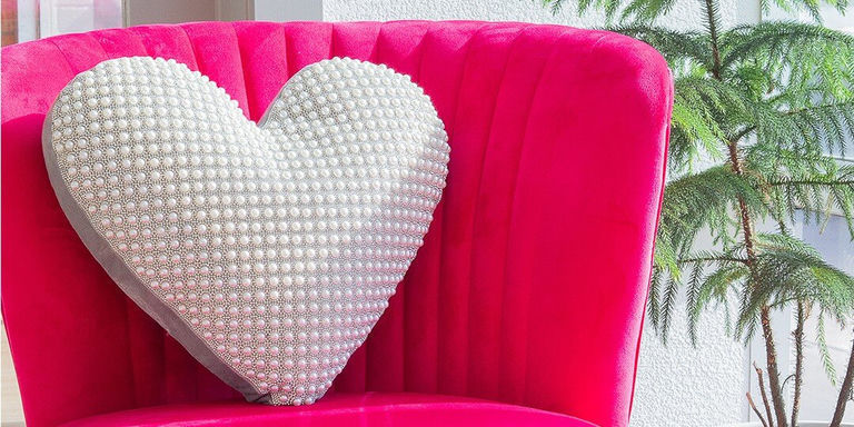 Silla de terciopelo rosa brillante con un gran cojín en forma de corazón cubierto de perlas blancas o diamantes de imitación, con planta de follaje verde visible al lado de la silla.