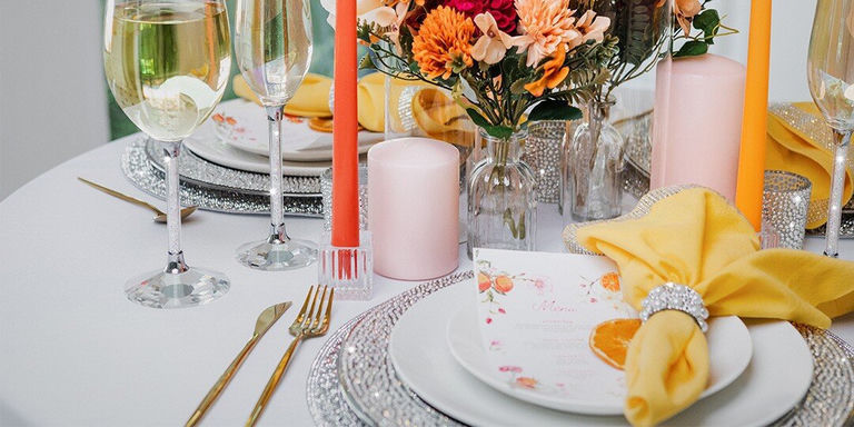 Une table élégante aux tonalités chaudes orange et jaune. Le couvert comprend des assiettes blanches à délicats motifs floraux, des couverts dorés, des verres à vin en cristal remplis de vin blanc et des serviettes en tissu jaune vif. Des bougies piliers orange dans des photophores en verre et un centre de table composé de fleurs orange et bordeaux dans des vases en verre transparent créent une atmosphère de repas d’automne festive. Des assiettes de présentation et des ronds de serviette scintillants ajoutent des touches glamour à cette table de saison.
