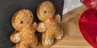 sparkly gingerbread man and woman holiday pillow styled on a rustic wooden floor beside a fireplace, a red tree skirt in the periphery