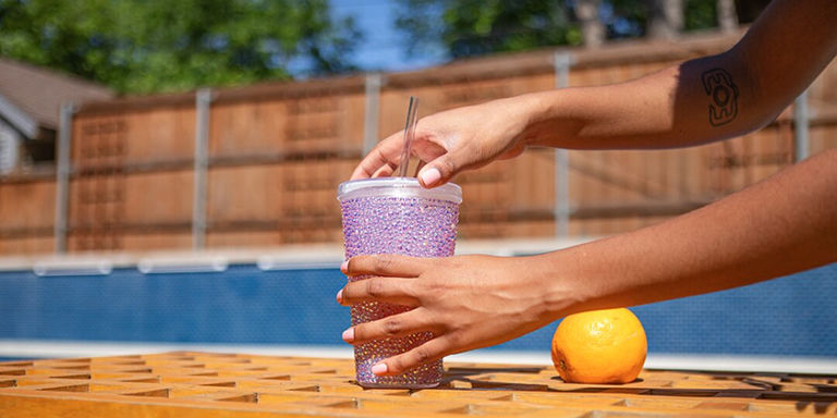 Gobelet en strass lavande tenu par des mains au bord de la piscine avec une orange