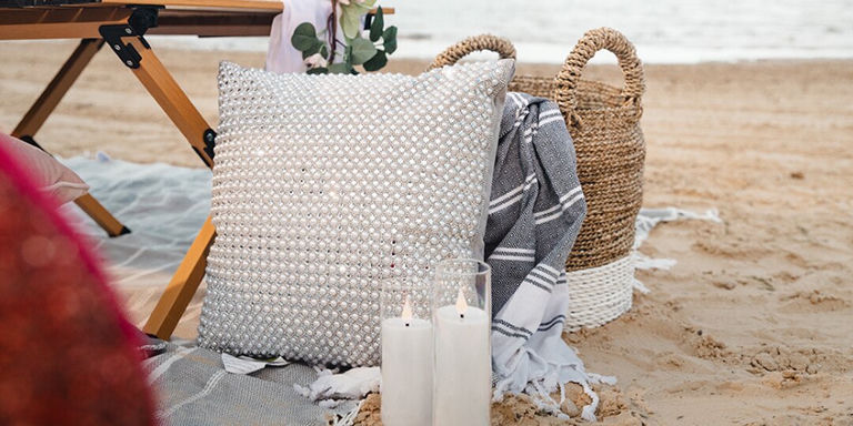 Installation de pique-nique sur la plage avec coussin décoratif en strass, bougies et panier tressé sur le sable