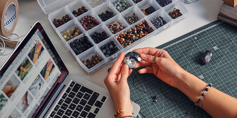 Hands working on rhinestone jewelry at a craft desk with gemstone storage box and laptop nearby