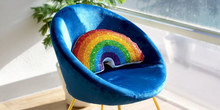 cojín de acento brillante arcoíris estilizado en una silla de terciopelo azul cerca de una ventana iluminada por el sol