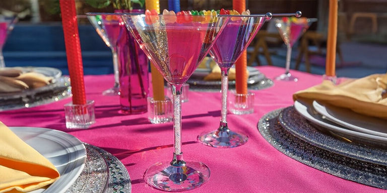 cócteles de orgullo coloridos servidos en copas de martini con strass con guarnición de gomitas arcoíris sobre mantel rosa brillante