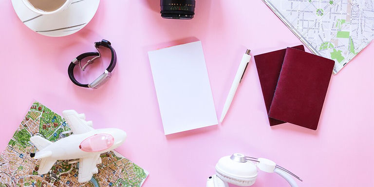 Flatlay of travel essentials including passports, maps, coffee, and camera on a pink background