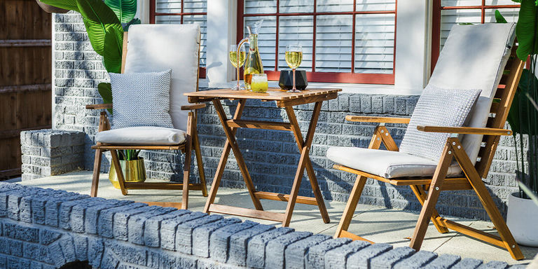 Patio exterior moderno con sillas de madera acolchadas blancas, mesa plegable de teca con copas de champán y plantas tropicales