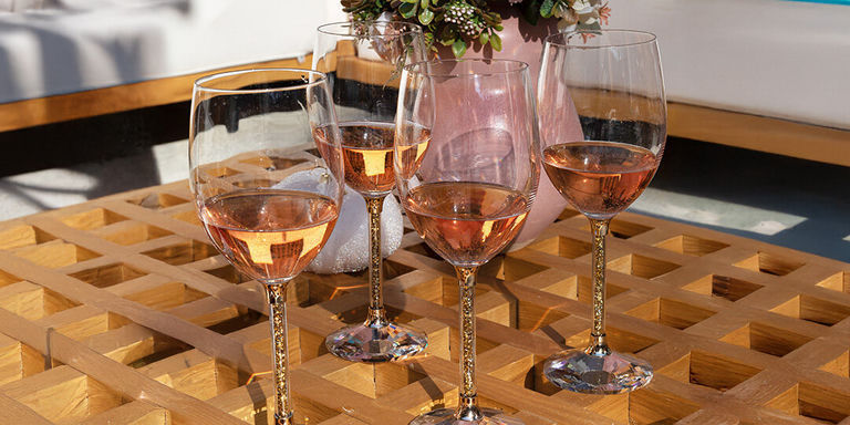 Collection de verres à vin aux pieds remplis de strass, garnis de vin rosé, disposés sur une surface en damier géométrique en bois. Différents formats de verres, flûtes à champagne et verres à vin sont présentés, avec de petits pots de succulentes visibles en arrière-plan.